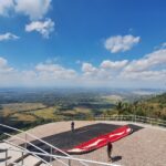 Group of people on a viewing platform overlooking a valley with fields below and a paragliding canopy on the ground nearby on a sunny day.