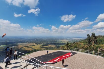 Group of people on a viewing platform overlooking a valley with fields below and a paragliding canopy on the ground nearby on a sunny day.