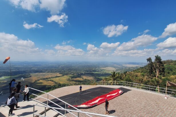 Group of people on a viewing platform overlooking a valley with fields below and a paragliding canopy on the ground nearby on a sunny day.