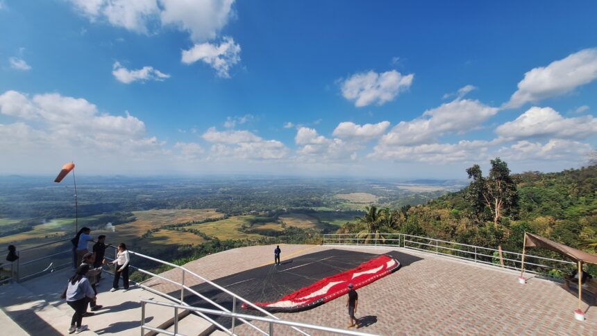 Group of people on a viewing platform overlooking a valley with fields below and a paragliding canopy on the ground nearby on a sunny day.