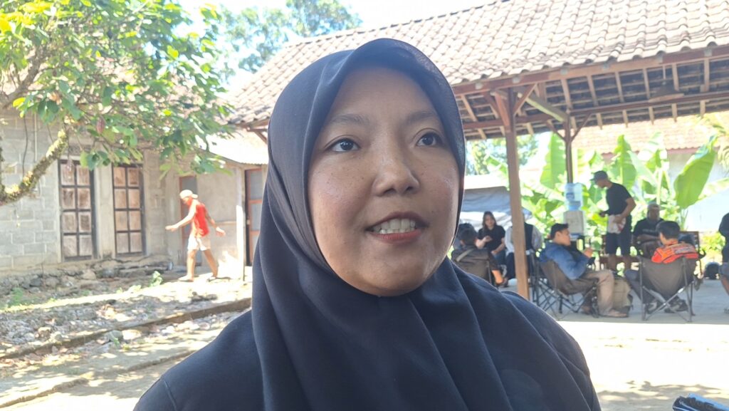 Close-up portrait of a woman wearing a dark hijab outdoors, with a group gathered under a wooden pavilion in the background.