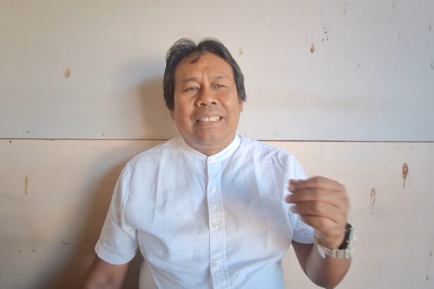 Man in a light blue shirt speaks to the camera, gesturing with his right hand against a plain wall.