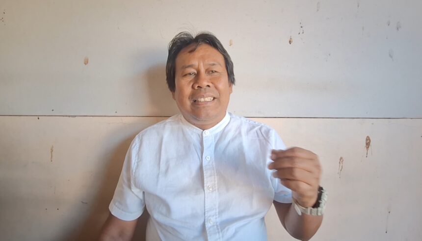 Man in a light blue shirt speaks to the camera, gesturing with his right hand against a plain wall.