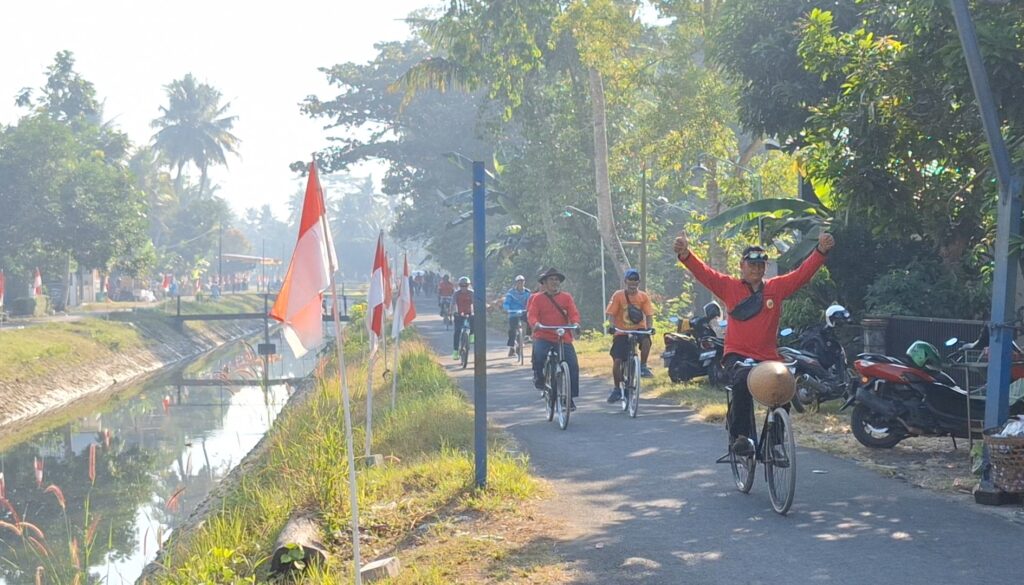 Screenshot_20260727_110838_Gallery - Mabur.co Cyclists ride along a sunny riverside path beside a canal; a man in red raises both arms in celebration with surrounding flags nearby.