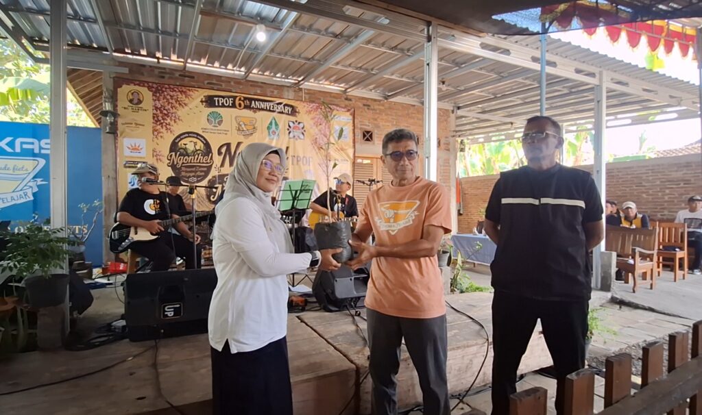 Screenshot_20260727_110940_Gallery - Mabur.co On a covered stage, a woman wearing a hijab hands a small potted plant to a man in an orange shirt during an anniversary event; musicians perform in the background and a man in black stands nearby.