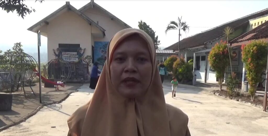 Woman in a tan hijab speaks to the camera in an outdoor courtyard with a playground and buildings in the background.