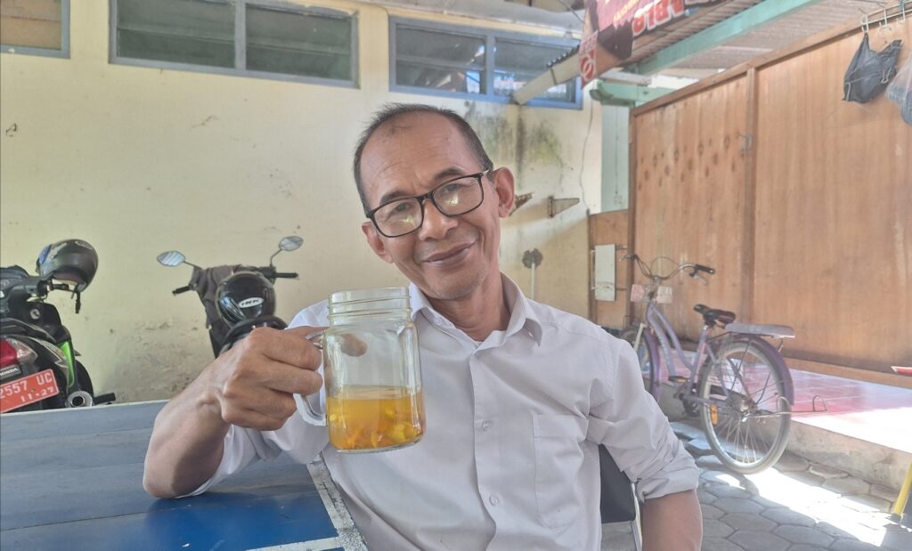 Screenshot_20260731_075024_Gallery - Mabur.co Man wearing glasses sits at a blue outdoor table, raising a mason jar of beer and smiling. Background shows motorcycles and a yellow wall.