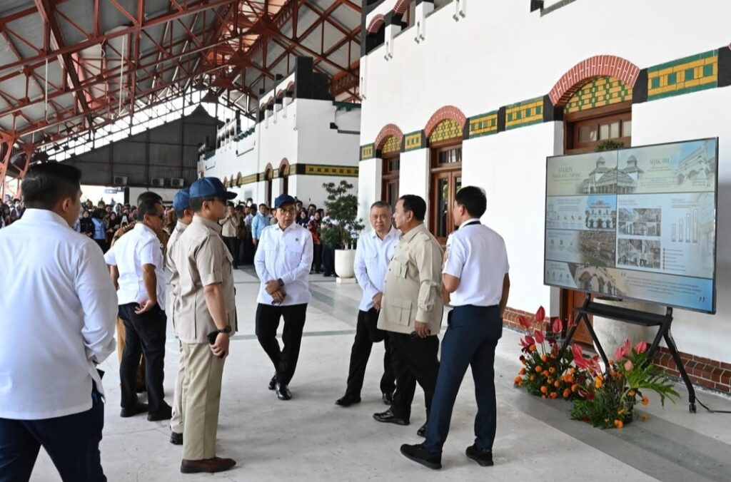 Group of officials and attendees in a covered hall, standing near a large display board with architectural drawings and photos, flowers nearby.