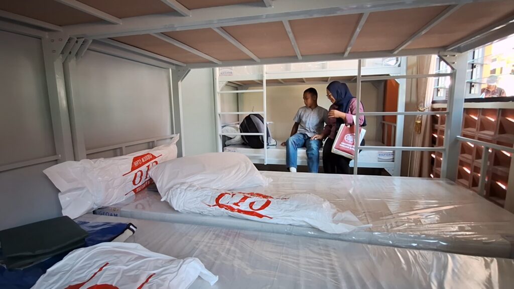 Inside a dorm-style room with metal bunk beds and plastic-wrapped mattresses; a boy sits on a lower bunk while a woman wearing a hijab shows a bag to him.