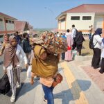 Crowd of people, many women in hijabs, gathered outside a building complex on a sunny day with orange-roofed structures nearby.