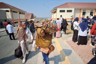 Crowd of people, many women in hijabs, gathered outside a building complex on a sunny day with orange-roofed structures nearby.