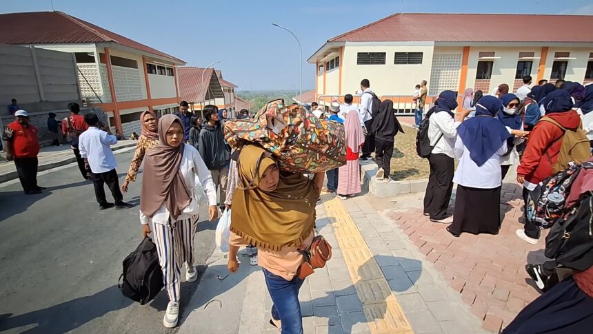 Crowd of people, many women in hijabs, gathered outside a building complex on a sunny day with orange-roofed structures nearby.
