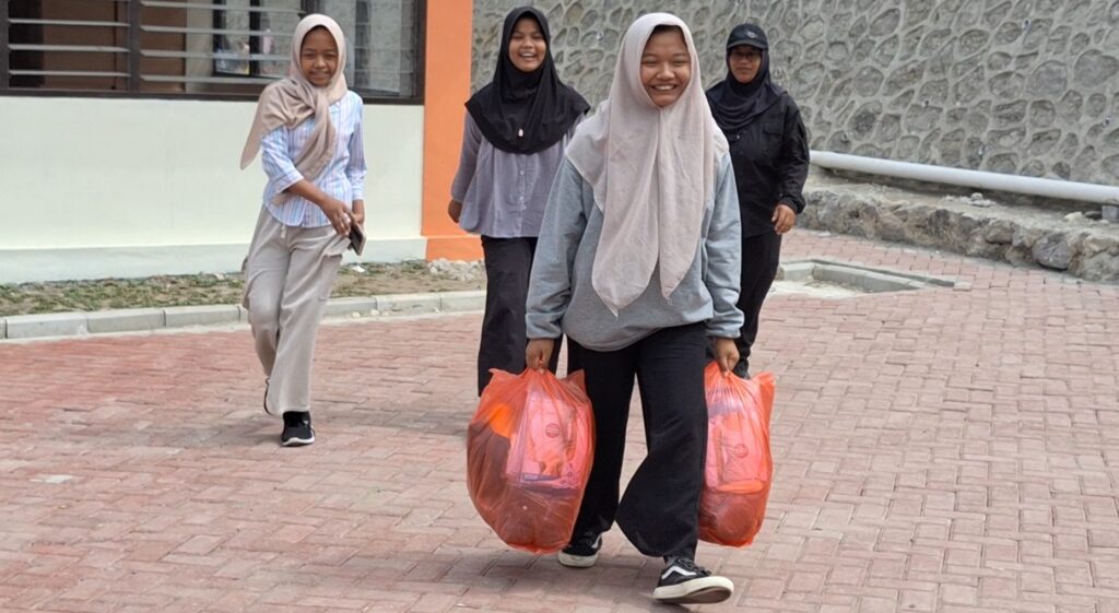 Four women wearing hijabs walk outdoors, smiling, two carrying bright orange bags.