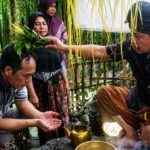 Man places a leafy sprig crown on a seated woman’s head as others observe, in an outdoor bamboo-walled area during a traditional blessing ritual.