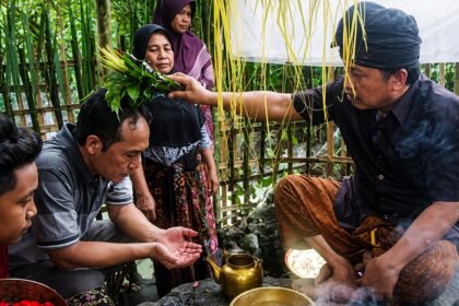 Man places a leafy sprig crown on a seated woman’s head as others observe, in an outdoor bamboo-walled area during a traditional blessing ritual.