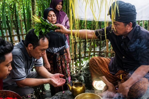 Man places a leafy sprig crown on a seated woman’s head as others observe, in an outdoor bamboo-walled area during a traditional blessing ritual.
