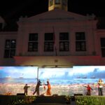 Nighttime stage performance in front of a white government building with 'GOUVERNEURSKANTOOR' on the facade and LED screens showing a seaside scene with ships.