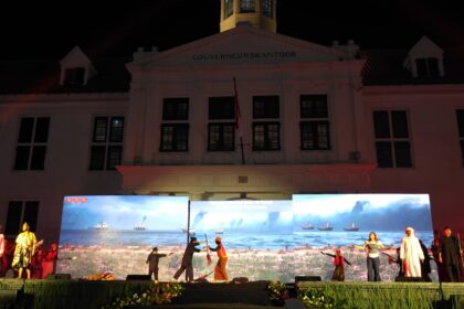 Nighttime stage performance in front of a white government building with 'GOUVERNEURSKANTOOR' on the facade and LED screens showing a seaside scene with ships.