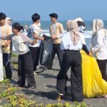 Group of volunteers on a beach collecting litter, passing filled bags toward a central pile by the sea.