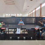 Wheelchair table tennis match in progress; two players in wheelchairs face off at a table, with a referee and a 4–6 scoreboard in the background.