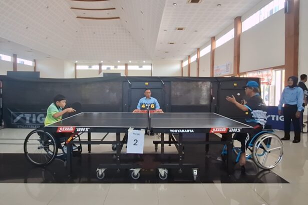 Wheelchair table tennis match in progress; two players in wheelchairs face off at a table, with a referee and a 4–6 scoreboard in the background.