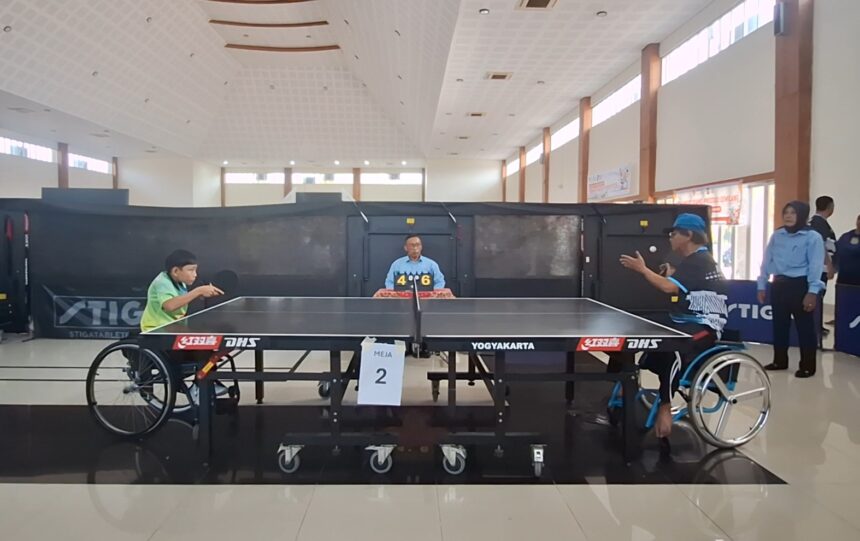 Wheelchair table tennis match in progress; two players in wheelchairs face off at a table, with a referee and a 4–6 scoreboard in the background.