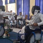Wheelchair fencers in full protective gear and masks compete in an indoor demo, with spectators watching behind blue curtains.