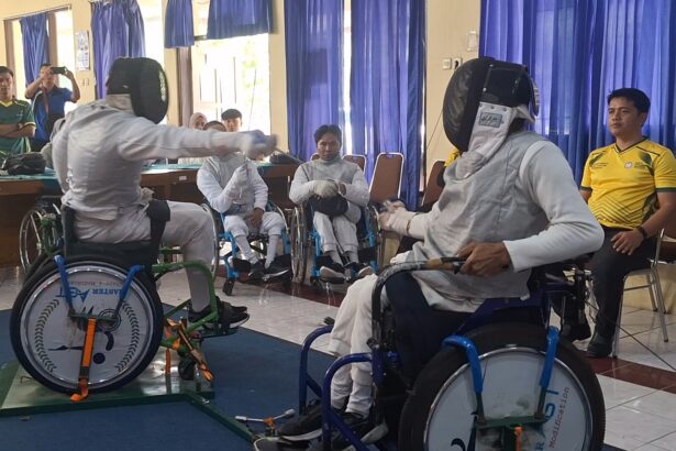 Wheelchair fencers in full protective gear and masks compete in an indoor demo, with spectators watching behind blue curtains.
