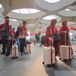 Group of travelers in red traditional outfits with rolling white suitcases in a modern airport terminal with a sculpted ceiling.