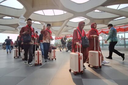 Group of travelers in red traditional outfits with rolling white suitcases in a modern airport terminal with a sculpted ceiling.