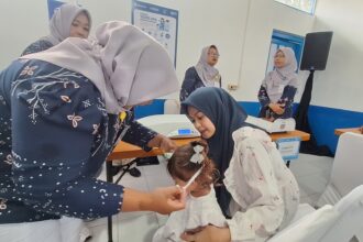 Women in hijabs assist a young girl in a clinic, performing a health check or procedure while others observe in the background.