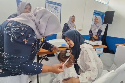 Women in hijabs assist a young girl in a clinic, performing a health check or procedure while others observe in the background.
