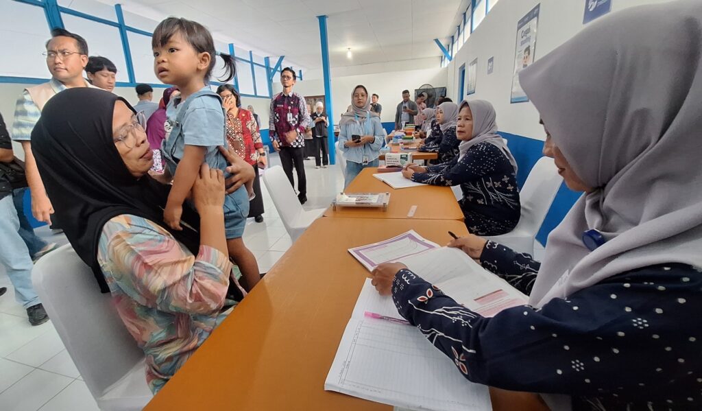 Screenshot_20260804_153047_Gallery - Mabur.co Woman in a black hijab holds a young child as staff at a desk fill out forms in a busy clinic or registration area.