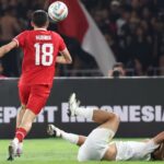Soccer player in a red jersey with the number 18 (Hubner) jumps to contest the ball as a white-clad opponent lies on the ground nearby on the pitch.