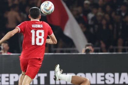 Soccer player in a red jersey with the number 18 (Hubner) jumps to contest the ball as a white-clad opponent lies on the ground nearby on the pitch.