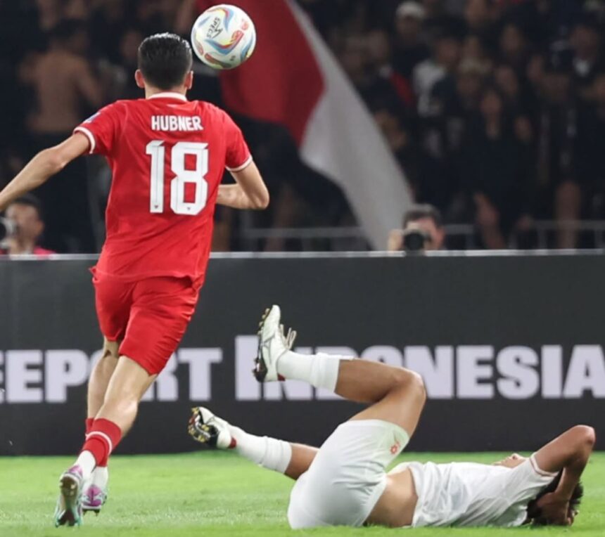 Soccer player in a red jersey with the number 18 (Hubner) jumps to contest the ball as a white-clad opponent lies on the ground nearby on the pitch.