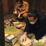 Two people wearing masks and gloves sort stacks of folders and envelopes on a green patterned mat inside a wooden room.