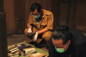 Two people wearing masks and gloves sort stacks of folders and envelopes on a green patterned mat inside a wooden room.