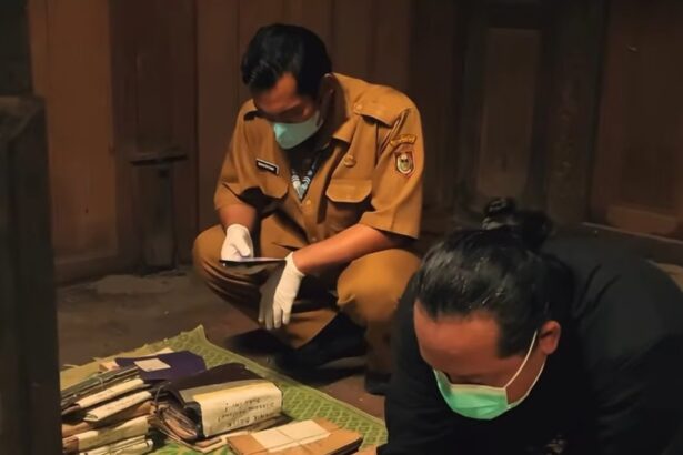 Two people wearing masks and gloves sort stacks of folders and envelopes on a green patterned mat inside a wooden room.