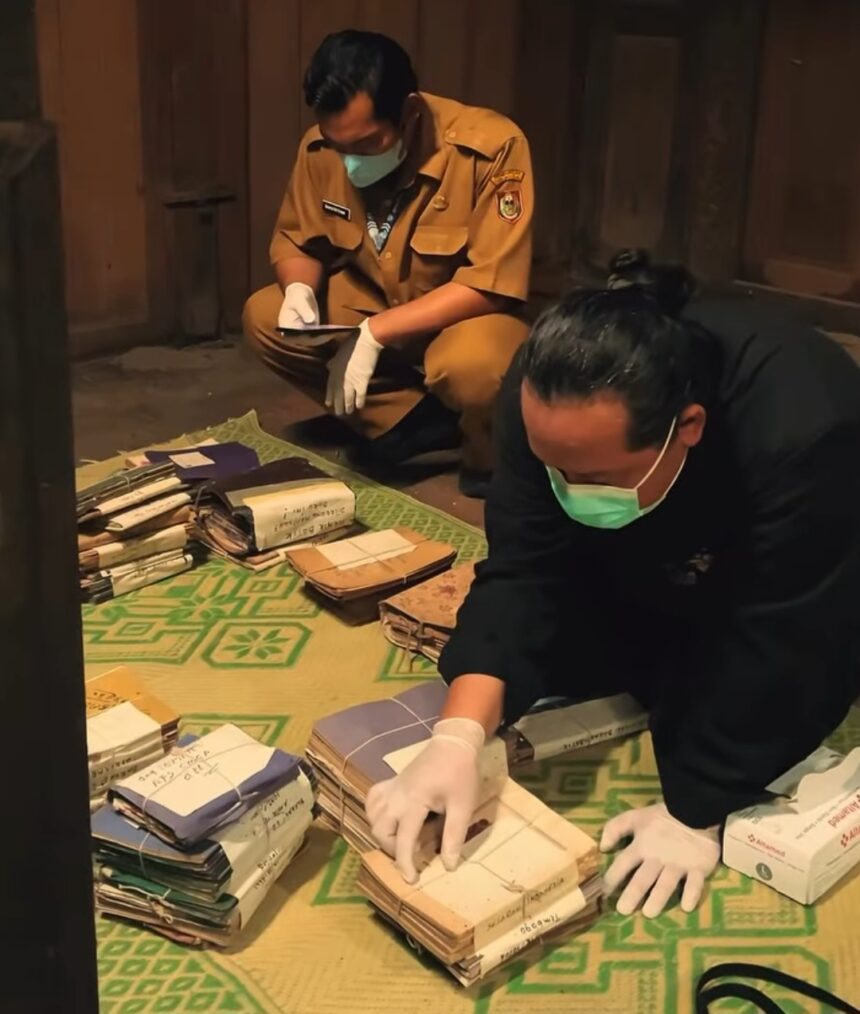 Two people wearing masks and gloves sort stacks of folders and envelopes on a green patterned mat inside a wooden room.