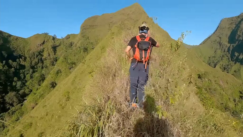 Hiker wearing an orange backpack and black shirt walks along a narrow grassy ridge with steep green slopes and a blue sky.