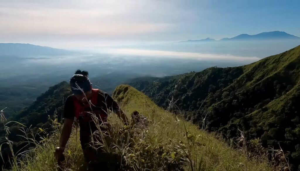 Hiker standing on a grassy ridge overlooking a misty valley and distant mountains at sunrise