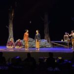 Actors in traditional costumes perform a forest-scene on a dark stage; two women stand center while others sit or pose around them.