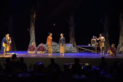 Actors in traditional costumes perform a forest-scene on a dark stage; two women stand center while others sit or pose around them.