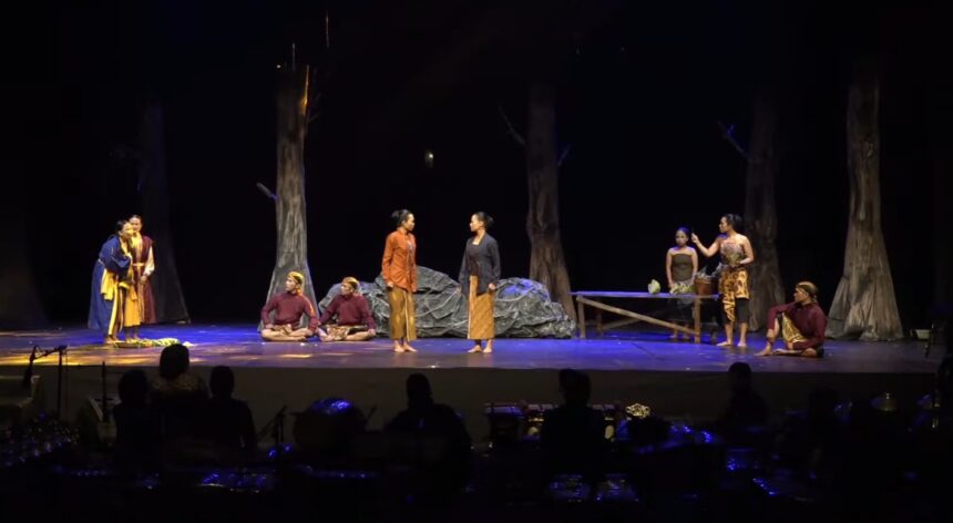 Actors in traditional costumes perform a forest-scene on a dark stage; two women stand center while others sit or pose around them.