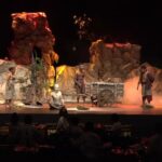 Stage scene from a play with actors in primitive clothing around a wooden cart on a rocky set with smoke effects, darkened audience area behind.
