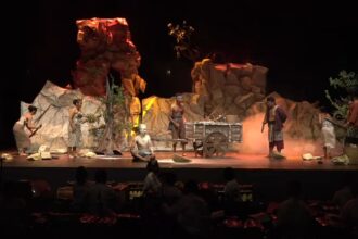 Stage scene from a play with actors in primitive clothing around a wooden cart on a rocky set with smoke effects, darkened audience area behind.