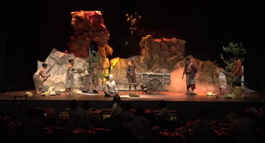 Stage scene from a play with actors in primitive clothing around a wooden cart on a rocky set with smoke effects, darkened audience area behind.