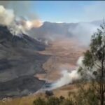 Volcanic eruption with lava flow and smoke rising from a rocky slope; scorched hillside and sparse trees in the foreground.