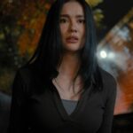 Young woman with long black hair in a dark top stands outdoors at night, illuminated by blurred city lights in the background.